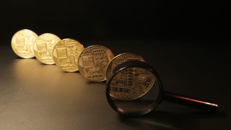 Fünf goldene Kryptowährungsmünzen, die in einer Reihe auf einer dunklen Fläche angeordnet sind, mit einer Lupe über der Münze, die dem Betrachter am nächsten ist, um das Konzept der Tokenisierung im digitalen Finanzwesen hervorzuheben.