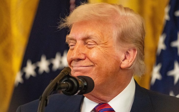 Ein Mann in Anzug und Krawatte steht lächelnd vor einem Mikrofon, während im Hintergrund US-Flaggen und Trump-Banner zu sehen sind.