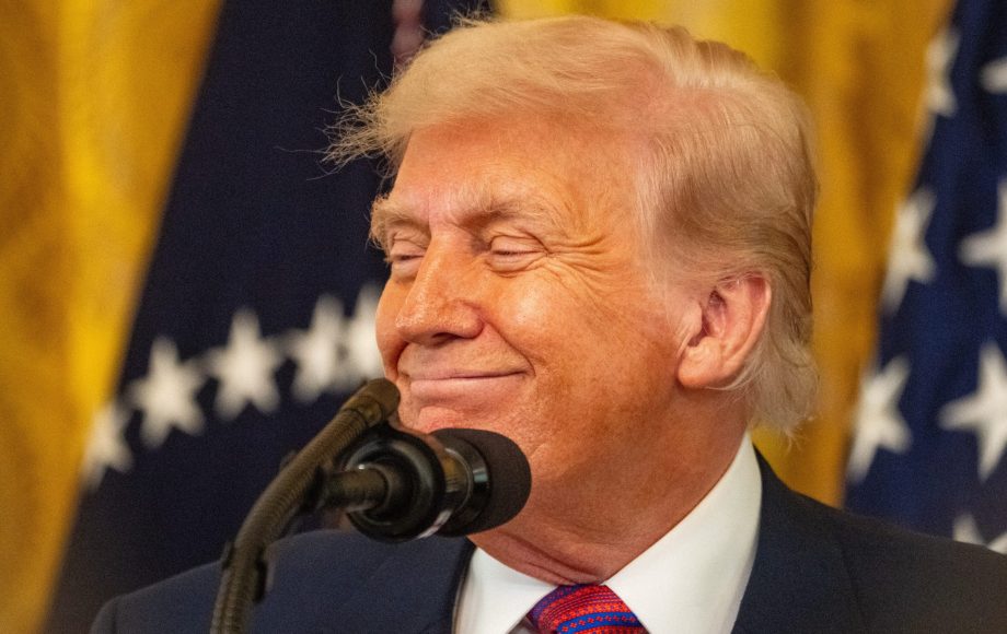 Ein Mann in Anzug und Krawatte steht lächelnd vor einem Mikrofon, während im Hintergrund US-Flaggen und Trump-Banner zu sehen sind.