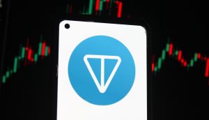 Ein Smartphone zeigt das Toncoin-Logo auf dem Bildschirm an, während im Hintergrund ein digitaler Candlestick-Chart von Toncoin mit roten und grünen Balken zu sehen ist.