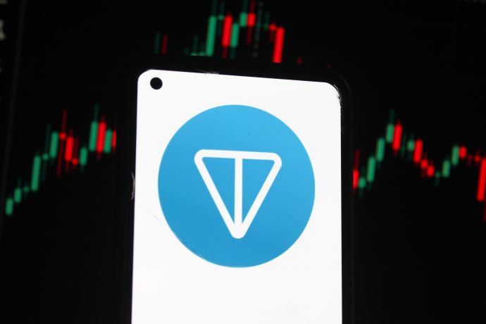 Ein Smartphone zeigt das Toncoin-Logo auf dem Bildschirm an, während im Hintergrund ein digitaler Candlestick-Chart von Toncoin mit roten und grünen Balken zu sehen ist.