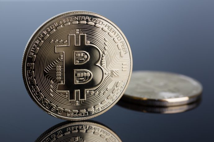 Eine Nahaufnahme einer physischen Bitcoin-Münze, die aufrecht steht, mit einer anderen Bitcoin, die flach im Hintergrund auf einer reflektierenden Oberfläche liegt.