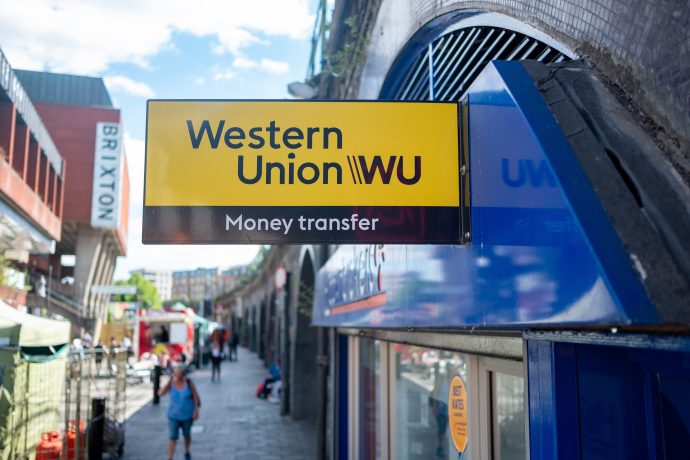 Ein Western-Union-Überweisungsschild ist über einem Schaufenster in einer städtischen Straßenszene mit Fußgängern und Marktständen angebracht und verdeutlicht den Kontrast zwischen traditionellen Überweisungen und neuen Optionen wie Stablecoins.