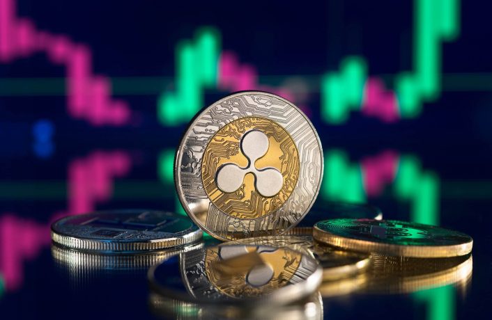 Mehrere XRP-Münzen sind auf einer spiegelnden Oberfläche gestapelt, vor einem unscharfen Hintergrund, der digitale Finanzcharts in Grün und Rosa anzeigt.