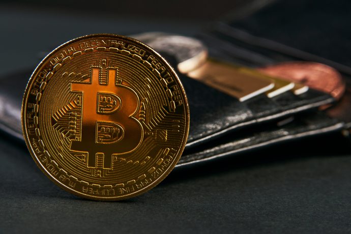 Eine goldene Bitcoin-Münze steht aufrecht vor einem schwarzen Lederportemonnaie, das die wachsende Überschneidung von Bitcoin und traditionellen Währungen mit sichtbaren Karten und Münzen im Hintergrund hervorhebt.