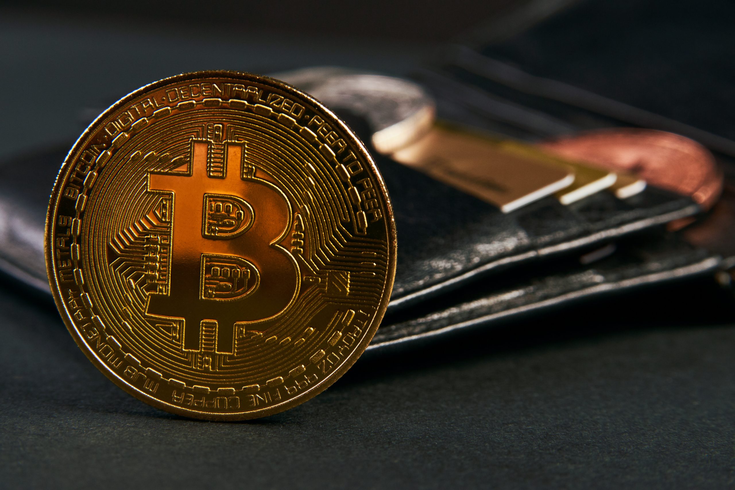 Eine goldene Bitcoin-Münze steht aufrecht vor einem schwarzen Lederportemonnaie, das die wachsende Überschneidung von Bitcoin und traditionellen Währungen mit sichtbaren Karten und Münzen im Hintergrund hervorhebt.