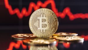 Eine Bitcoin-Münze steht aufrecht zwischen anderen Münzen. Im unscharfen Hintergrund sind eine rote, abwärts gerichtete Kurve und US-Dollar-Symbole zu erkennen.