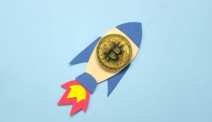 Eine goldene Bitcoin-Münze auf einem Papierausschnitt einer Rakete vor einem hellblauen Hintergrund symbolisiert eine optimistische Bitcoin-Prognose.