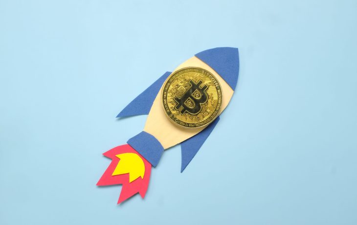 Eine goldene Bitcoin-Münze auf einem Papierausschnitt einer Rakete vor einem hellblauen Hintergrund symbolisiert eine optimistische Bitcoin-Prognose.