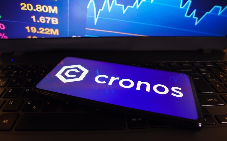 Ein Smartphone mit dem Cronos-Logo liegt auf einer Laptop-Tastatur, während auf dem Laptop-Bildschirm im Hintergrund Kursziele und Börsenzahlen erscheinen.