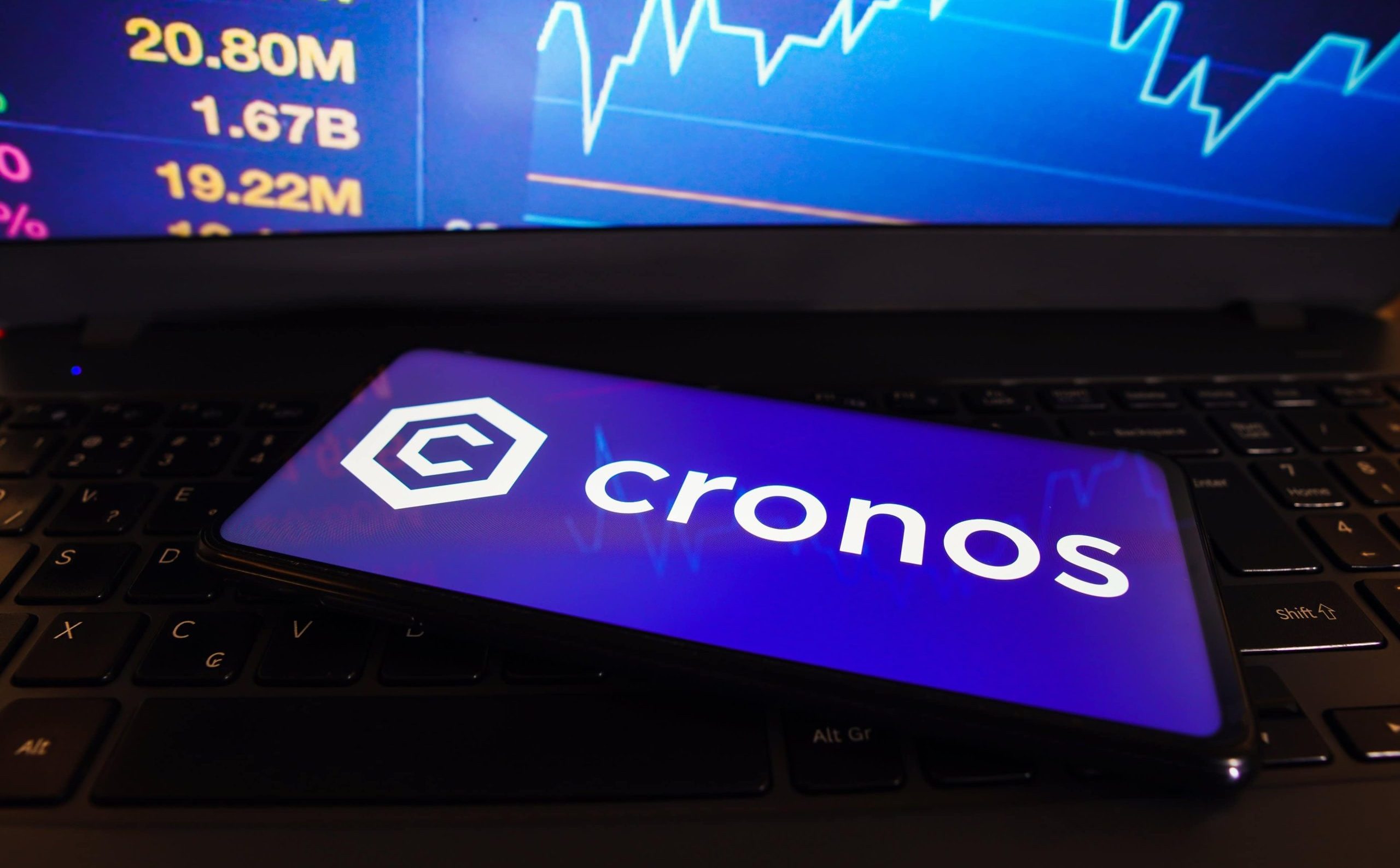 Ein Smartphone mit dem Cronos-Logo liegt auf einer Laptop-Tastatur, während auf dem Laptop-Bildschirm im Hintergrund Kursziele und Börsenzahlen erscheinen.