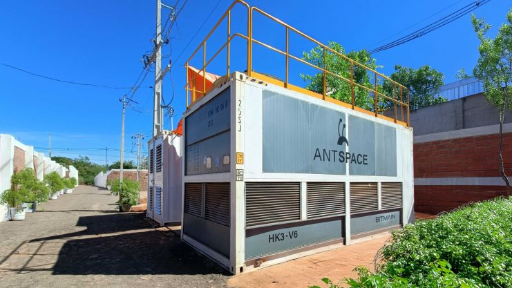 Ein großer weißer Metall-Bergbaucontainer mit der Aufschrift ANTSPACE und BITMAIN steht unter freiem Himmel an einer Ziegelmauer.