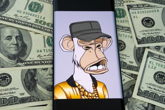 Auf einem Smartphone ist ein Bored Ape Yacht Club NFT zu sehen, der einen Hut und eine Goldkette trägt und von verstreuten Hundert-Dollar-Scheinen umgeben ist - perfekt für jeden versierten Anleger.