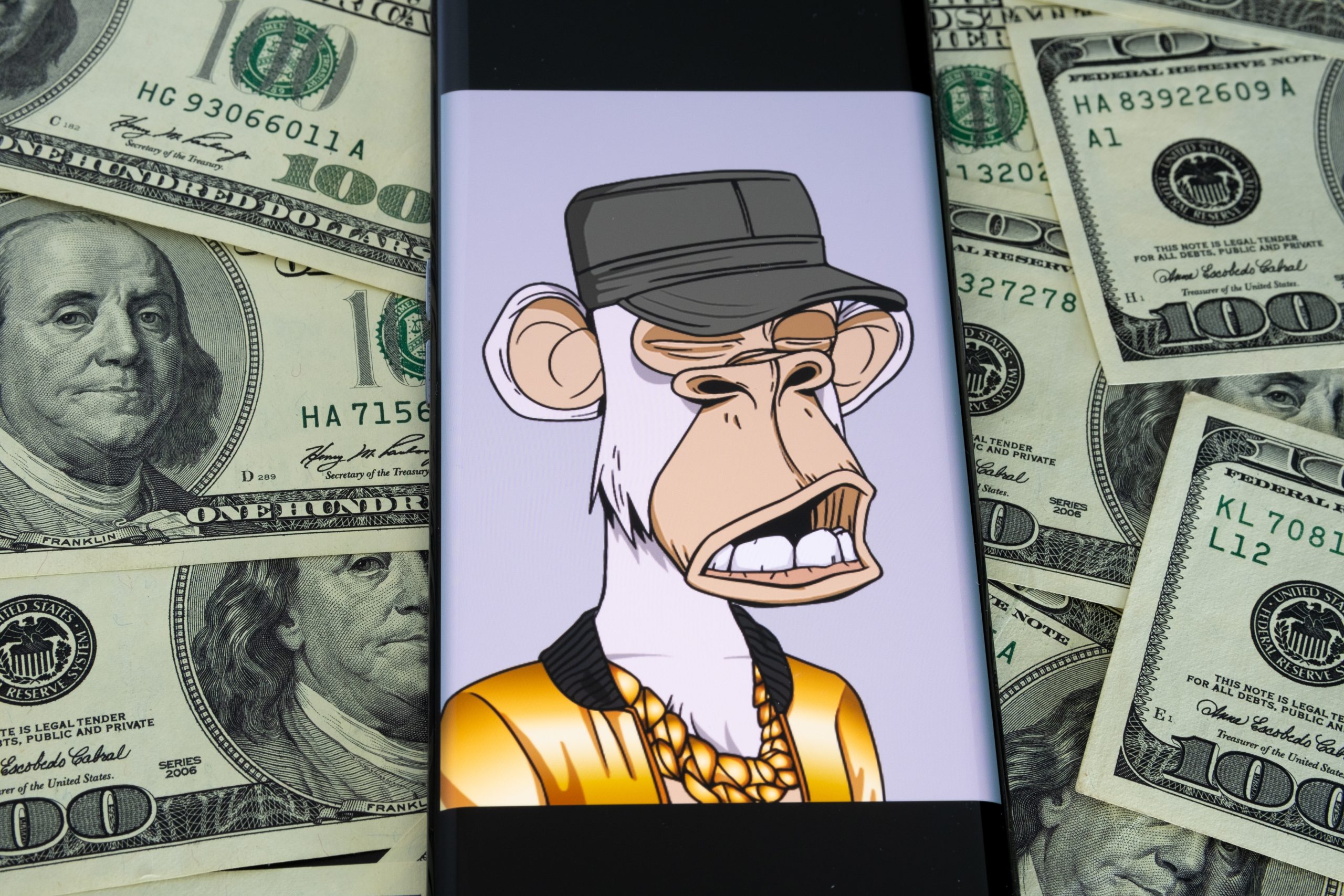 Auf einem Smartphone ist ein Bored Ape Yacht Club NFT zu sehen, der einen Hut und eine Goldkette trägt und von verstreuten Hundert-Dollar-Scheinen umgeben ist - perfekt für jeden versierten Anleger.