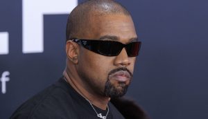 Kanye West Ye mit rasiertem Kopf, Bart und Schnurrbart trägt eine dunkle Sonnenbrille und ein schwarzes Hemd und steht vor einem dunklen Hintergrund mit weißen Buchstaben, der stark an Kanye West erinnert.