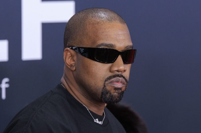 Kanye West Ye mit rasiertem Kopf, Bart und Schnurrbart trägt eine dunkle Sonnenbrille und ein schwarzes Hemd und steht vor einem dunklen Hintergrund mit weißen Buchstaben, der stark an Kanye West erinnert.