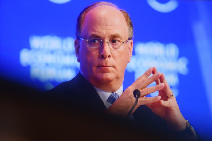 Larry Fink der CEO von BlackRock in Anzug und Krawatte sitzt mit verschränkten Fingern da und schaut zur Seite. Der blaue Hintergrund zeigt verschwommene Texte und Logos, die auf Finanzthemen wie Bitcoin-ETFs oder die Stärke des USD hinweisen.