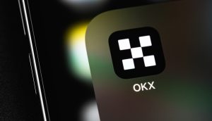 Nahaufnahme eines Smartphone-Bildschirms mit dem OKX-App-Symbol, das einen schwarzen Hintergrund mit einem weiß karierten OKX-Logo und dem Text "OKX" darunter zeigt.