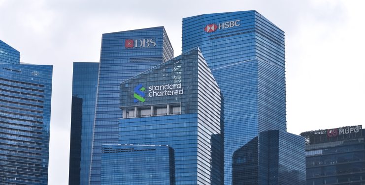 Eine Ansammlung hoher Bürogebäude aus blauem Glas, darunter die Logos der Ripple-Partner DBS, HSBC, Standard Chartered und MUFG, heben sich vom bewölkten Himmel ab.