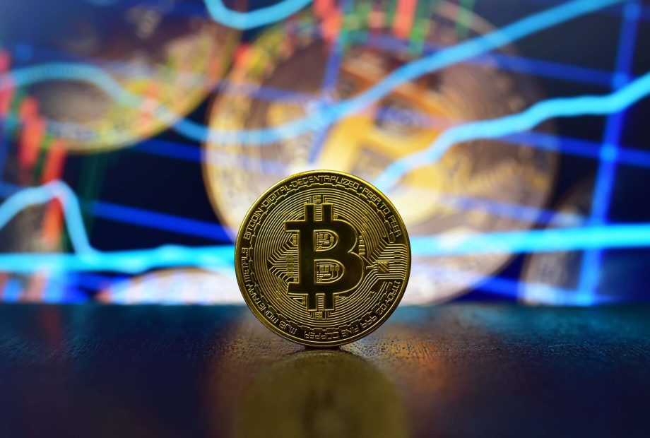 Eine goldene Bitcoin-Münze steht aufrecht auf einem Tisch, während digitale Charts und Finanzdiagramme die Bitcoin-Trends im verschwommenen Hintergrund hervorheben.