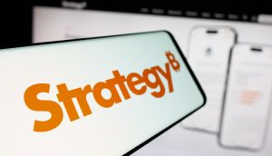 Ein Smartphone zeigt das Wort Strategy mit einem Bitcoin-Symbol vor einem unscharfen Computerbildschirm, der die Benutzeroberfläche einer mobilen Anwendung zeigt.