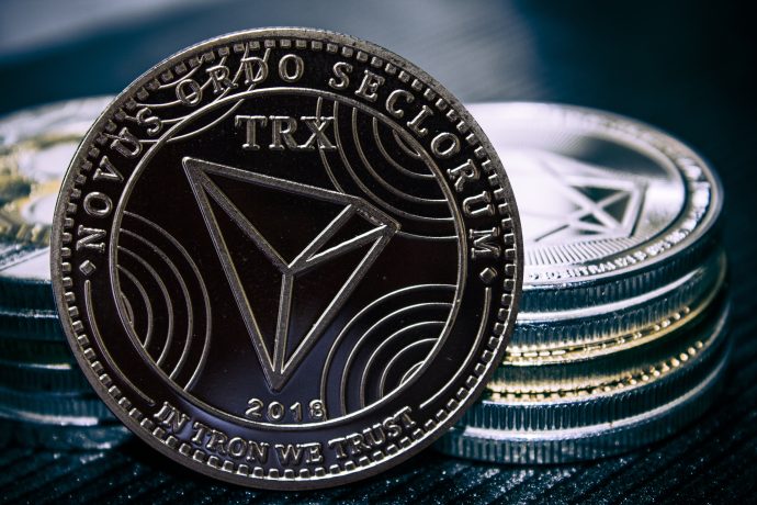 Eine Nahaufnahme einer Tron (TRX) Kryptowährungsmünze steht aufrecht vor gestapelten Münzen und zeigt detaillierte Gravuren und den Text NOVUS ORDO SECLORUM und IN TRON WE TRUST.