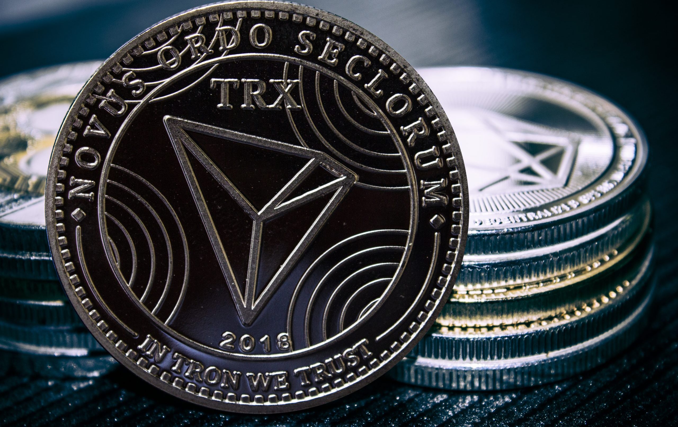 Eine Nahaufnahme einer Tron (TRX) Kryptowährungsmünze steht aufrecht vor gestapelten Münzen und zeigt detaillierte Gravuren und den Text NOVUS ORDO SECLORUM und IN TRON WE TRUST.