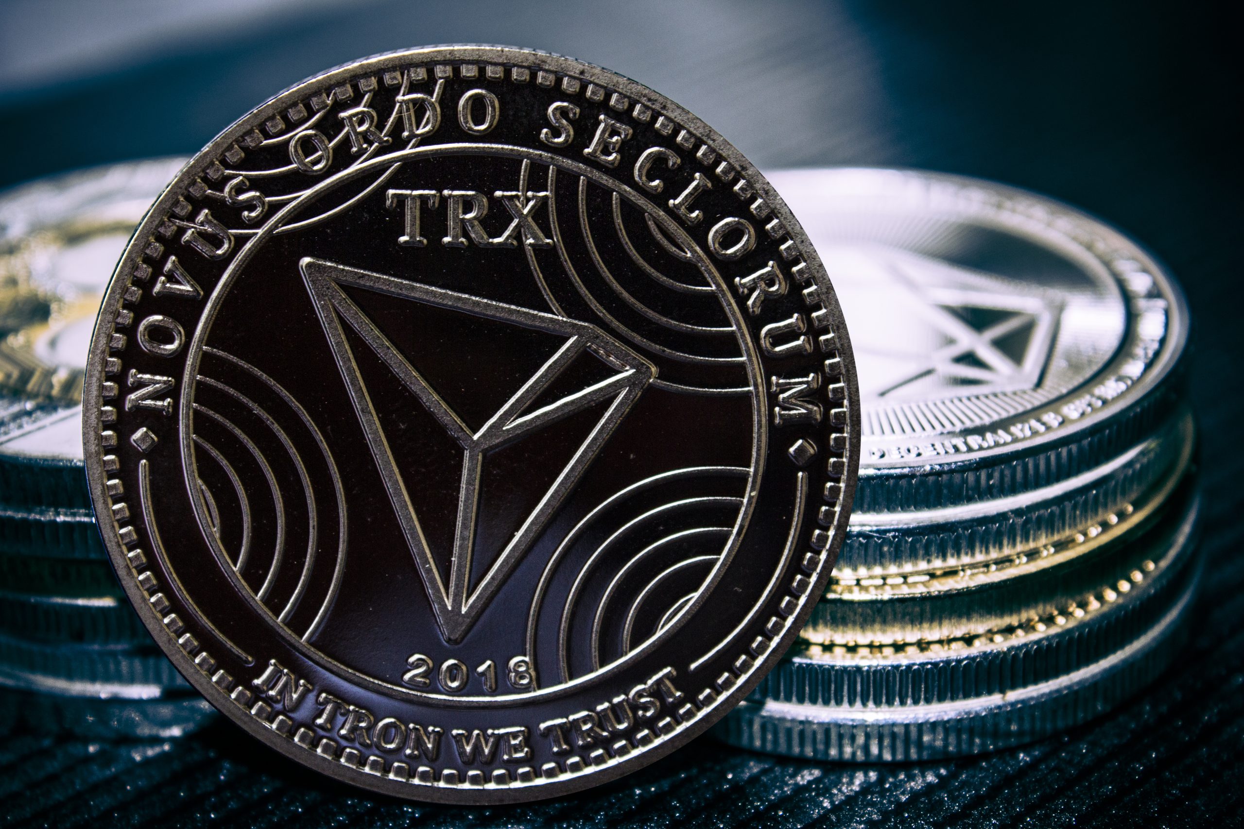Eine Nahaufnahme einer Tron (TRX) Kryptowährungsmünze steht aufrecht vor gestapelten Münzen und zeigt detaillierte Gravuren und den Text NOVUS ORDO SECLORUM und IN TRON WE TRUST.
