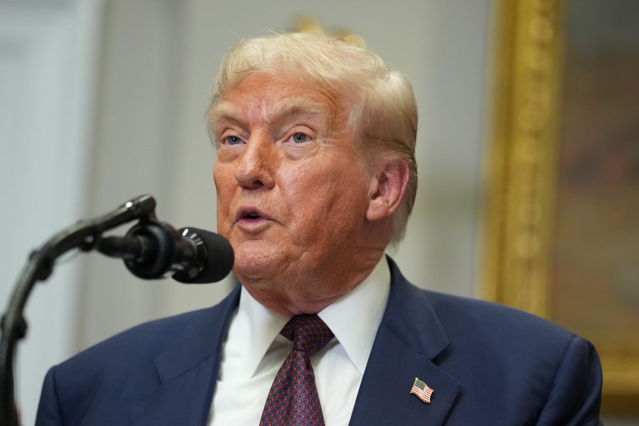 Donald Trump in einem marineblauen Anzug und roter Krawatte, der Trump ähnelt, spricht in ein Mikrofon an einem Podium und trägt eine Anstecknadel mit der amerikanischen Flagge an seiner Jacke.