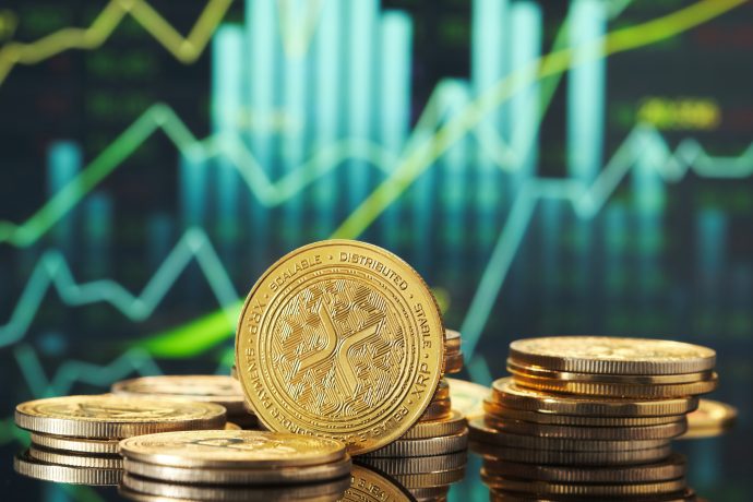 Im Mittelpunkt stehen Stapel goldfarbener Kryptowährungsmünzen, darunter XRP, mit einem digitalen Finanzdiagramm, das im unscharfen Hintergrund steigende Graphen anzeigt.