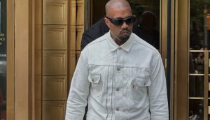 Kanye West mit einer dunklen Sonnenbrille und einer weißen Jeansjacke geht vor einem Gebäude mit goldfarbenen Türen.