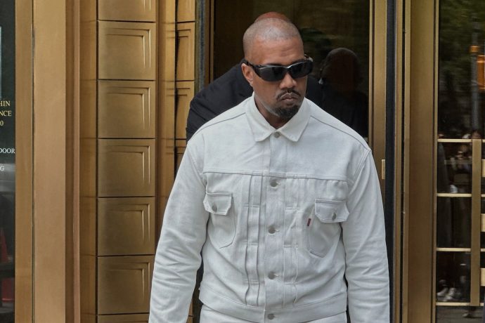 Kanye West mit einer dunklen Sonnenbrille und einer weißen Jeansjacke geht vor einem Gebäude mit goldfarbenen Türen.