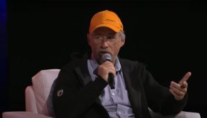 Ein älterer Mann, der eine gelbe McLaren-Mütze und eine Brille trägt, diskutiert über Bitcoin, während er in ein Mikrofon spricht. Er sitzt auf einem weißen Stuhl vor einem dunklen Hintergrund.