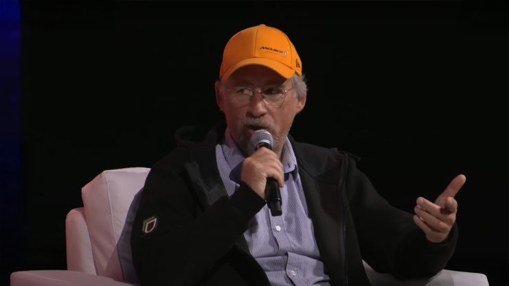 Ein älterer Mann, der eine gelbe McLaren-Mütze und eine Brille trägt, diskutiert über Bitcoin, während er in ein Mikrofon spricht. Er sitzt auf einem weißen Stuhl vor einem dunklen Hintergrund.