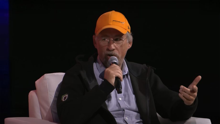 Ein älterer Mann, der eine gelbe McLaren-Mütze und eine Brille trägt, diskutiert über Bitcoin, während er in ein Mikrofon spricht. Er sitzt auf einem weißen Stuhl vor einem dunklen Hintergrund.