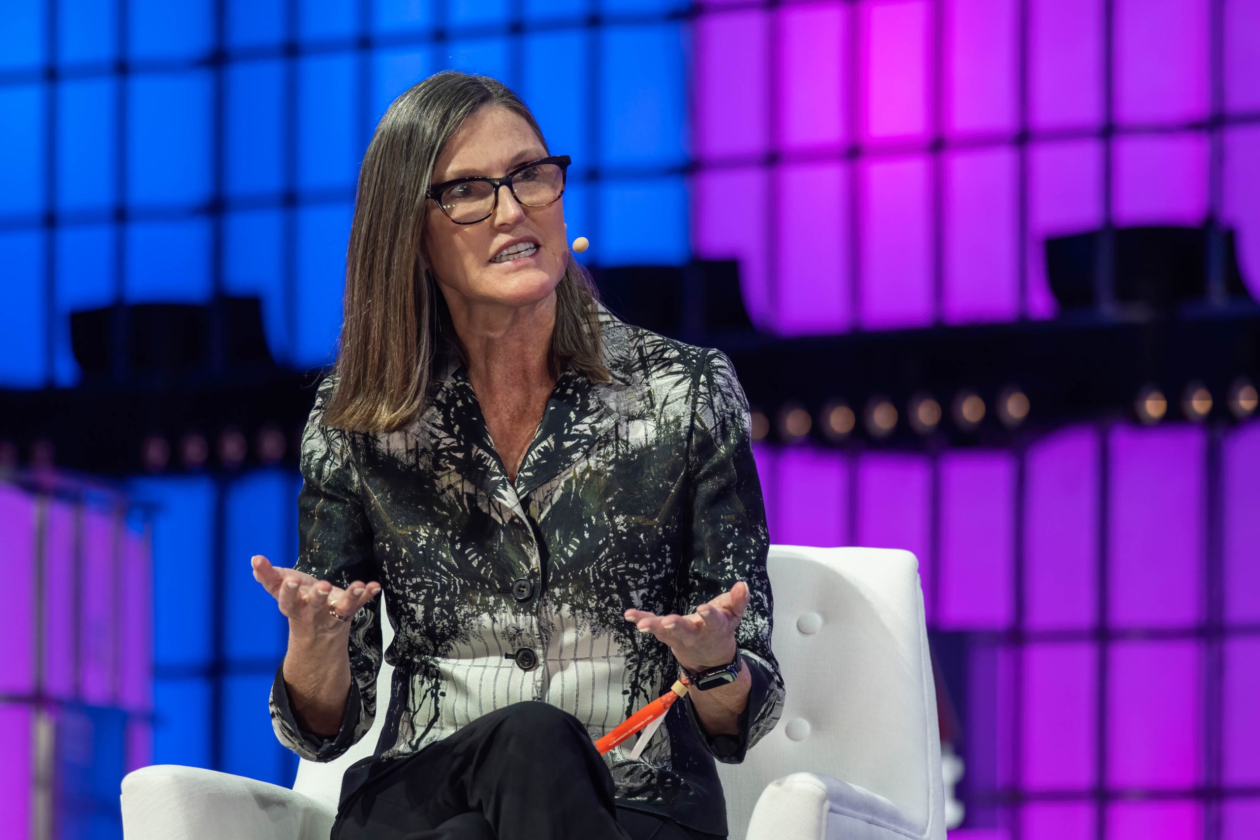 Cathie Wood sitzt auf einem weißen Stuhl und gestikuliert mit ihren Händen, während sie spricht; bunte blaue und violette Lichter füllen den Hintergrund.