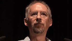 Ein Mann mittleren Alters mit Brille, kurzen grauen Haaren und Spitzbart blickt vor einem dunklen Hintergrund nach vorne, wobei sich das sanfte Leuchten eines Bitcoin-Symbols in seinen Gläsern spiegelt.