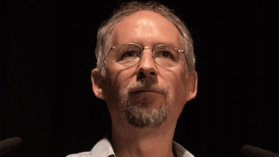 Ein Mann mittleren Alters mit Brille, kurzen grauen Haaren und Spitzbart blickt vor einem dunklen Hintergrund nach vorne, wobei sich das sanfte Leuchten eines Bitcoin-Symbols in seinen Gläsern spiegelt.