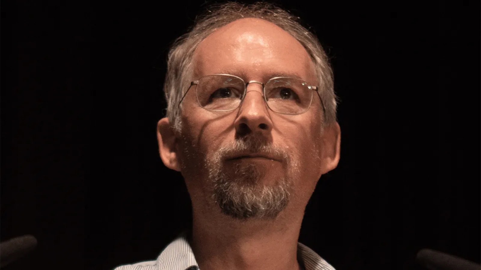 Ein Mann mittleren Alters mit Brille, kurzen grauen Haaren und Spitzbart blickt vor einem dunklen Hintergrund nach vorne, wobei sich das sanfte Leuchten eines Bitcoin-Symbols in seinen Gläsern spiegelt.