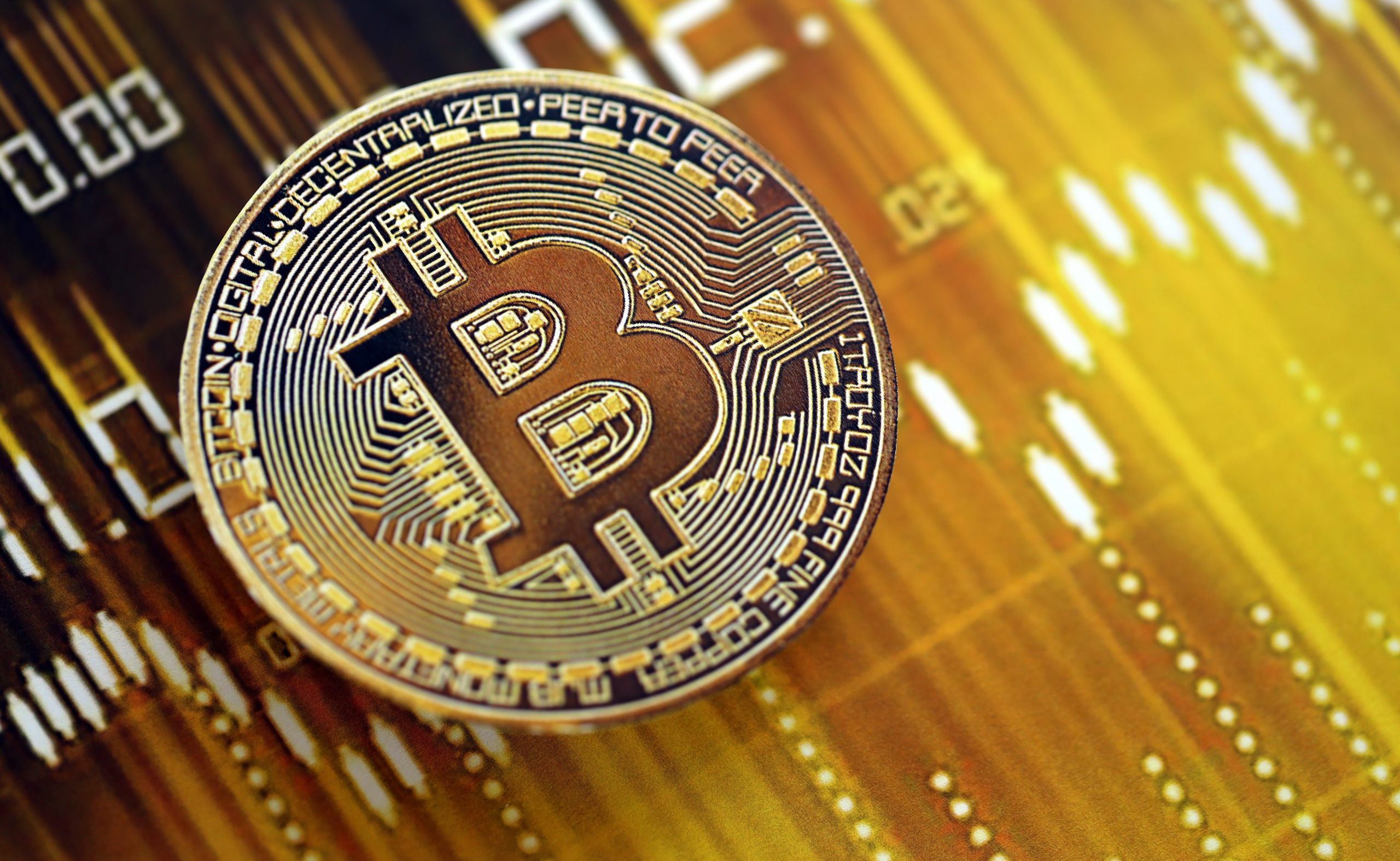 Eine physische Bitcoin-Münze wird vor einem unscharfen Hintergrund digitaler Finanzdaten angezeigt, die mit Gold- und Gelbtönen unterlegt sind.