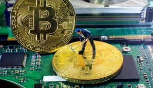 Eine kleine Figur eines Arbeiters steht auf einer großen Bitcoin-Münze auf einem Computer-Motherboard, ein weiterer Bitcoin steht aufrecht neben ihm und symbolisiert die Arbeit, die hinter dem Mining von Kryptowährungen steckt.