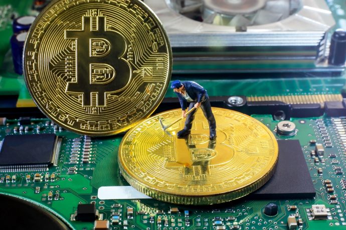 Eine kleine Figur eines Arbeiters steht auf einer großen Bitcoin-Münze auf einem Computer-Motherboard, ein weiterer Bitcoin steht aufrecht neben ihm und symbolisiert die Arbeit, die hinter dem Mining von Kryptowährungen steckt.