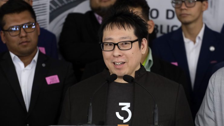 Ein Mann mit Brille und schwarzer Jacke spricht an einem Podium mit Mikrofonen über Bitcoin, umgeben von Menschen in Geschäftskleidung.