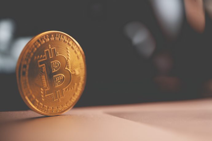 Eine Nahaufnahme einer einzelnen goldfarbenen Bitcoin, die aufrecht auf einer flachen Oberfläche steht und das ikonische Symbol vor einem dunklen, verschwommenen Hintergrund zeigt.