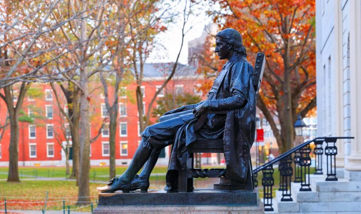Bronzestatue eines sitzenden Mannes auf einem Sockel vor einem Gebäude, mit herbstlichen Bäumen, roten Backsteinstrukturen und einem dezenten Bitcoin-Motiv im Hintergrund.