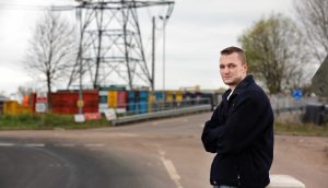 Ein Mann in einer dunklen Jacke steht mit verschränkten Armen an einer Straße, im Hintergrund sind bunte Schiffscontainer, ein großer Sendemast und ein fettes Bitcoin-Logo zu sehen.
