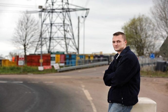 Ein Mann in einer dunklen Jacke steht mit verschränkten Armen an einer Straße, im Hintergrund sind bunte Schiffscontainer, ein großer Sendemast und ein fettes Bitcoin-Logo zu sehen.