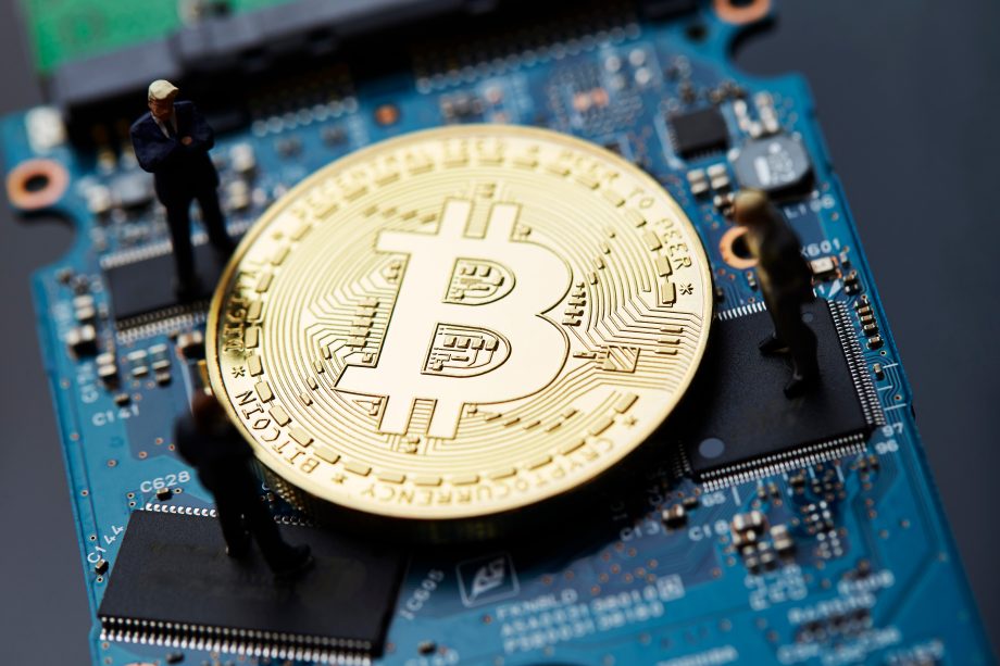 Eine große goldene Bitcoin-Münze ruht auf einer Computerplatine, in deren Nähe drei Miniaturfiguren von Menschen stehen, die den globalen Einfluss der Bitcoin-Technologie symbolisieren.