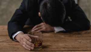 Eine Person im Anzug sitzt mit gesenktem Kopf an einem Holztisch, in der einen Hand ein Glas mit braunem Schnaps, scheinbar beunruhigt über den jüngsten Bitcoin-Absturz.