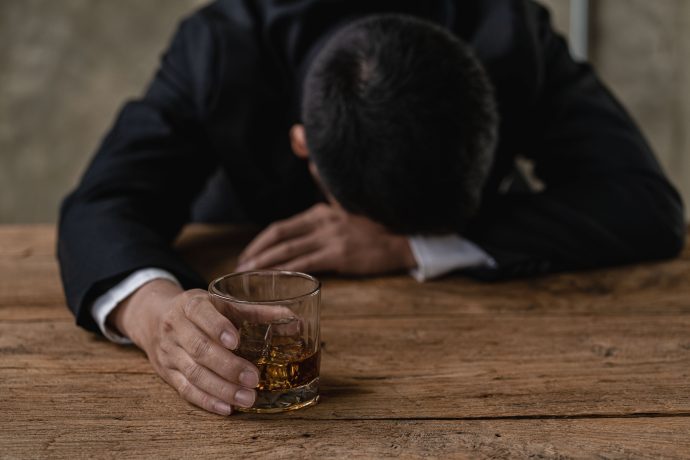 Eine Person im Anzug sitzt mit gesenktem Kopf an einem Holztisch, in der einen Hand ein Glas mit braunem Schnaps, scheinbar beunruhigt über den jüngsten Bitcoin-Absturz.